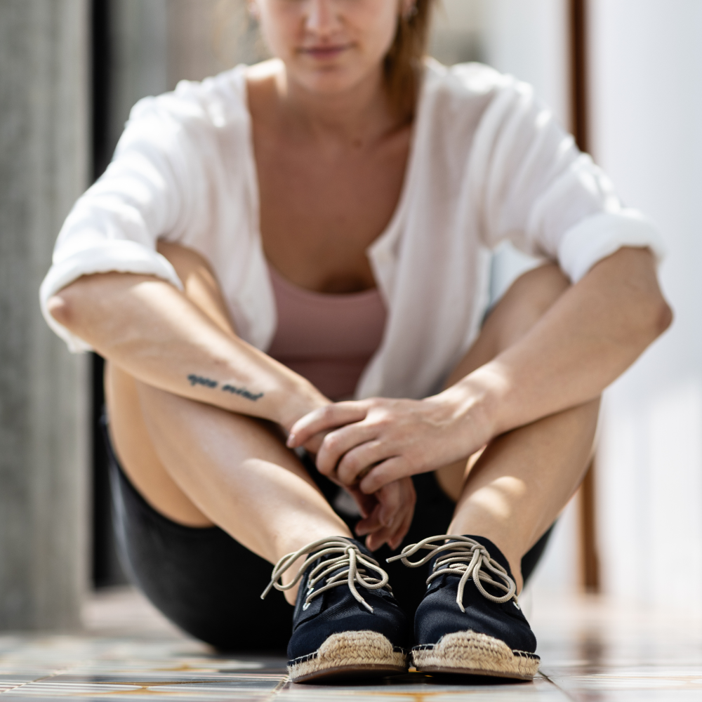 Woman sitting on a wooden floor wearing black espadrilles with beige soles.