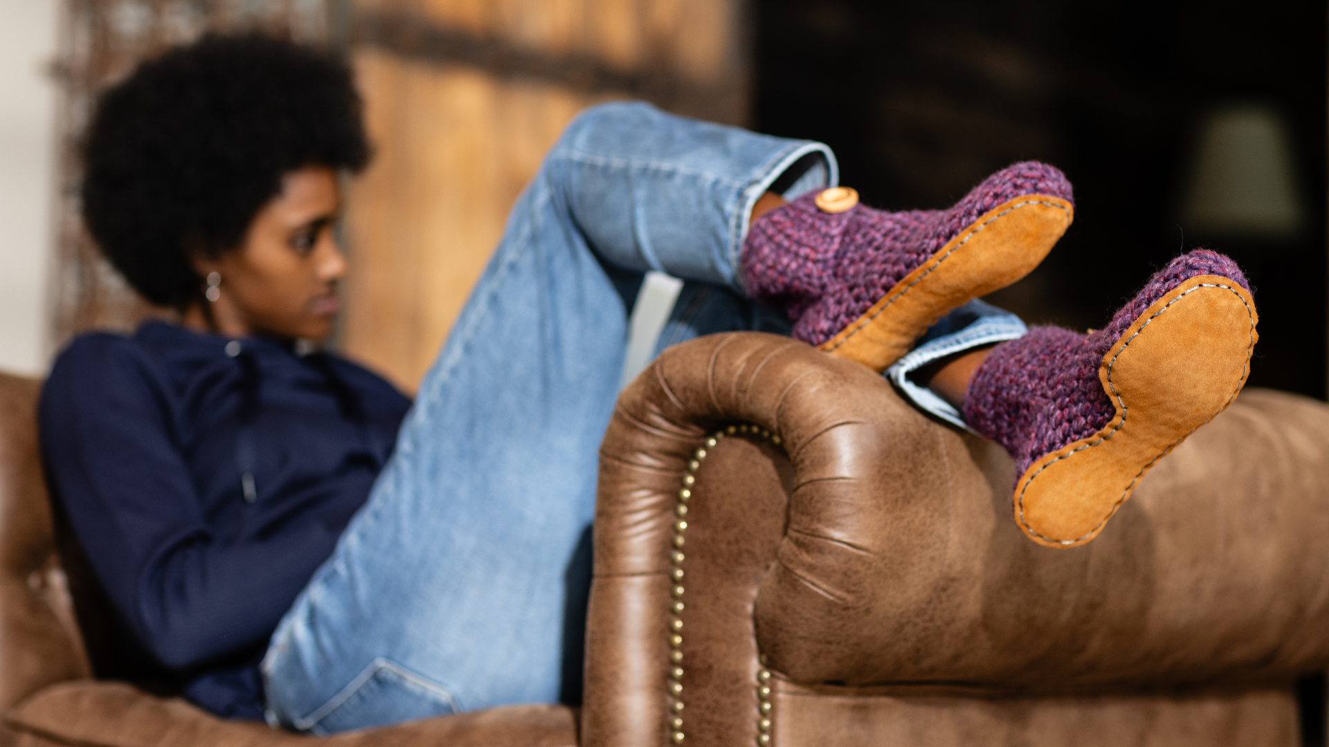 Person sitting on a brown leather couch with feet propped up wearing purple  slippers with leather soles.