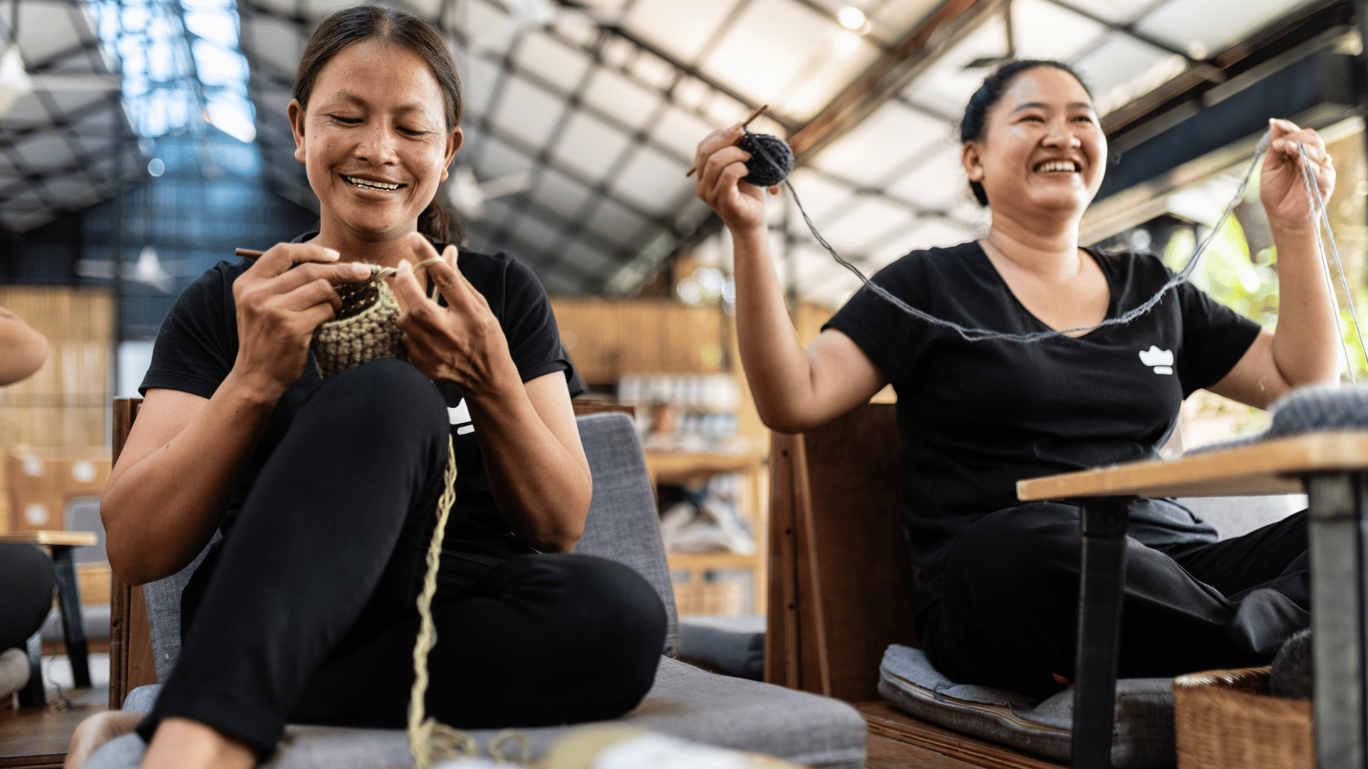 Two women sitting on chairs, one knitting and the other holding yarn, in a casual indoor setting.